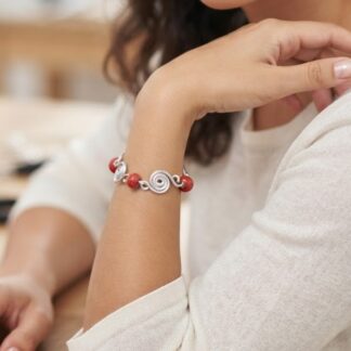 1791825146701 - Zanghi collection Close-up of a woman's wrist wearing a silver spiral-link bracelet with red beads on a light sweater, resting on a wooden table.