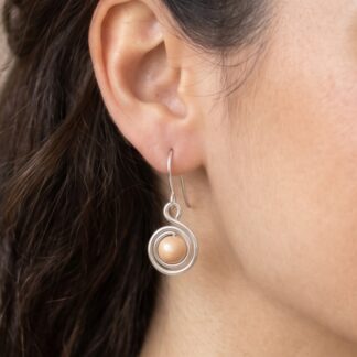 Close-up of a woman's left ear wearing a silver concentric-ring pendant earring with a peach bead at the center.