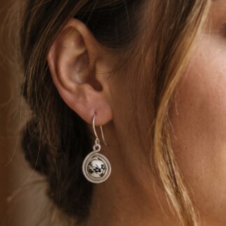 Close-up of a woman's ear wearing a silver hoop earring with a circular pendant featuring a white bead and black floral design.