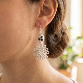 Close-up of a woman's ear wearing a dangling silver earring with a white bead featuring a black floral pattern.