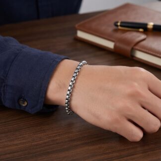 1792227446617 - Zanghi collection Forearm resting on a dark wooden desk, wearing a silver bracelet, with a brown leather notebook and black-gold pen in the background