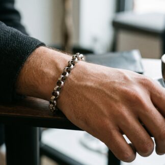 1792474565481 - Zanghi collection Close-up of a forearm resting on a table, wearing a silver chain bracelet, with a cup of coffee blurred in the background.