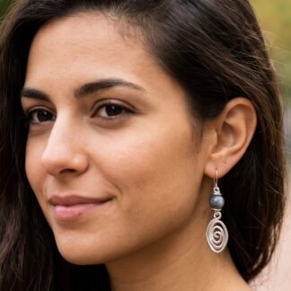 Close-up of a woman with dark hair wearing a silver spiral-eared dangling necklace-style earring with a gray bead, outdoors