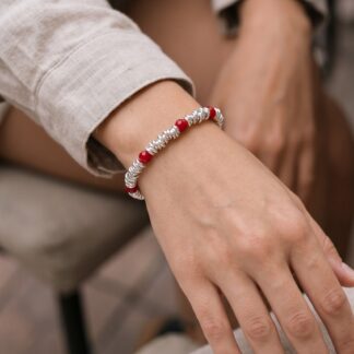1792573433877 - Zanghi collection Close-up of a wrist wearing a silver and red beaded bracelet, with a light beige rolled-up sleeve nearby.