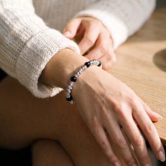 1792573708039 - Zanghi collection Close-up of a person resting their hands on their knee, wearing a cream knit sweater and a silver coil bracelet with black beads.