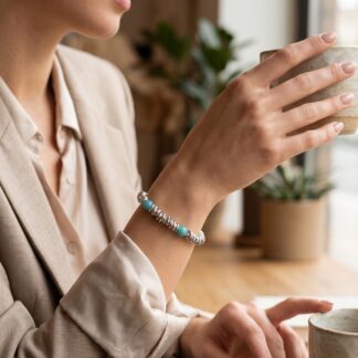 1792573953299 - Zanghi collection Close-up of a woman's hand holding a speckled ceramic coffee cup on a saucer by a window, wearing a beige blazer and turquoise bracelet.