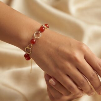 Bracelet on wrist with red beads and linked gold rings against a silky beige background.