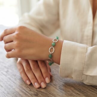 Close-up of a person's hands resting on a wooden surface, wearing a beaded bracelet with green beads and silver circular links on the wrist.