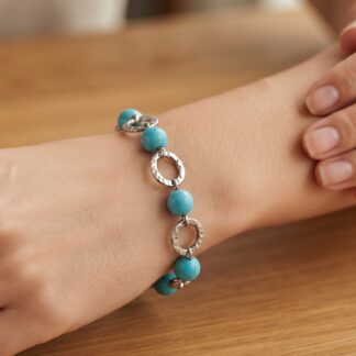 Close-up of a wrist wearing a turquoise bead bracelet with hammered silver rings on a wooden table surface.