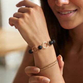 Close-up of a woman's wrist wearing a black lava bead bracelet with gold interlocking rings and delicate gold chains.
