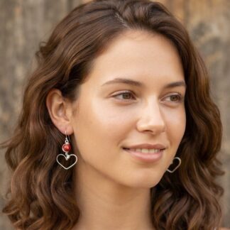 Close-up of a smiling woman with wavy brown hair, wearing silver heart earrings with a red bead outdoors.