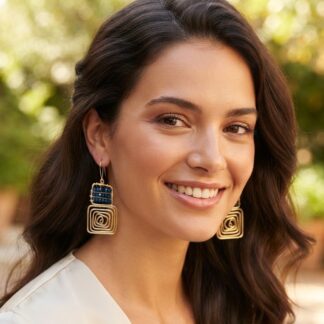 Close-up of a smiling woman with dark hair wearing large gold spiral earrings and a blue beaded accent, outdoors.