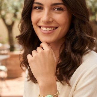 Woman smiling warmly outdoors, resting chin on hand, wearing a light sweater and a bracelet with a green bead.