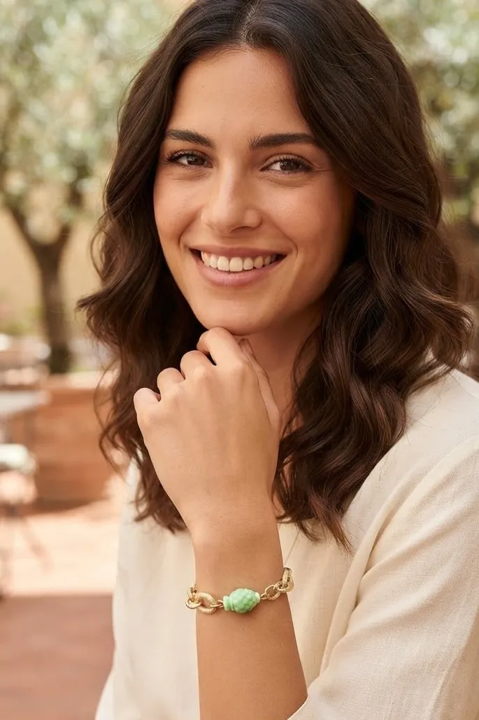 1791807681760 - Zanghi collection Woman smiling warmly outdoors, resting chin on hand, wearing a light sweater and a bracelet with a green bead.