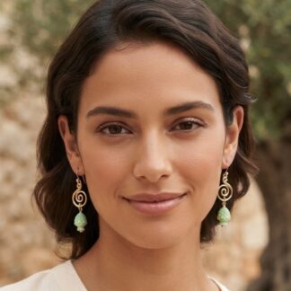 Close-up portrait of a woman with short dark hair, wearing gold spiral earrings with green beads, outdoors with a stone wall background.