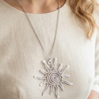Close-up of a silver sunburst pendant necklace with a spiraled center resting on a beige linen top. A silver sun pendant on a chain worn over a light cream blouse, shown from the chest up. Silver sun-shaped pendant with a spiral center on a thin chain against a pale beige fabric. Necklace featuring a sunburst pendant with winding lines, displayed on a light-colored top. Pendant: sun design with a spiral core, hanging from a delicate chain over a cream-colored garment.