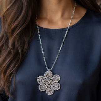 Woman wearing a silver chain necklace with a large flower-shaped pendant featuring intricate spiral metalwork against a dark blue blouse.